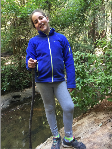 Emily Mitnick standing confidently on a rock with a walking stick during a hike along a riverside trail.